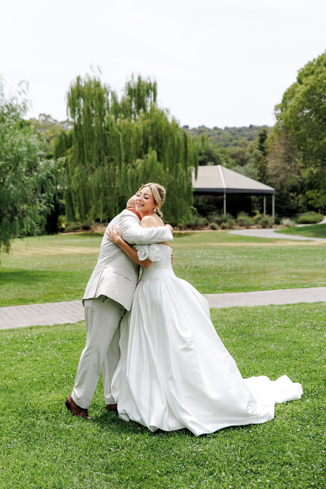 Bride and groom share a romantic embrace on lush green lawn, bride wearing elegant white gown with train.
