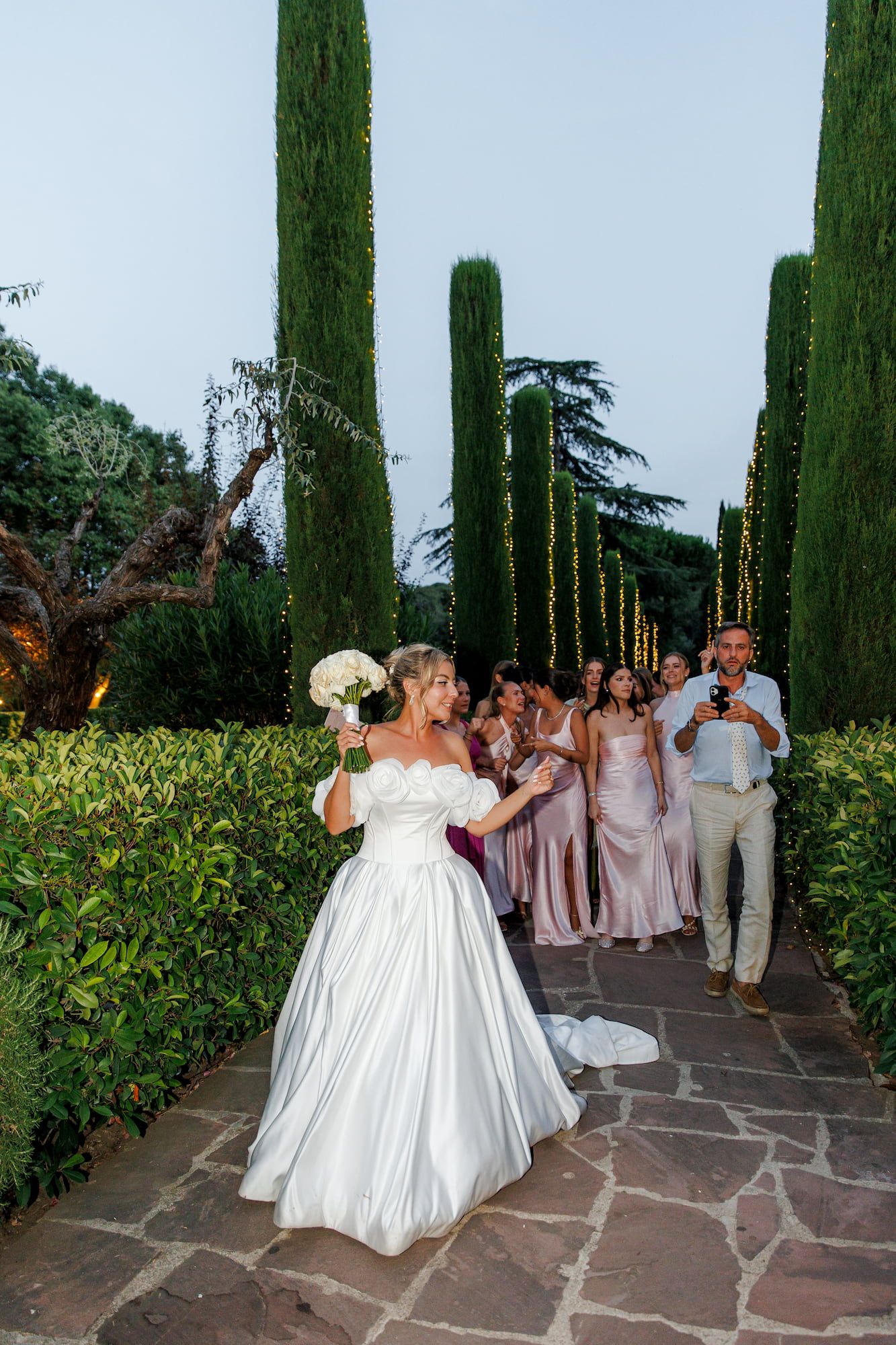 Bride in white gown tosses bouquet to bridesmaids in pink dresses at outdoor wedding venue with tall cypress trees.