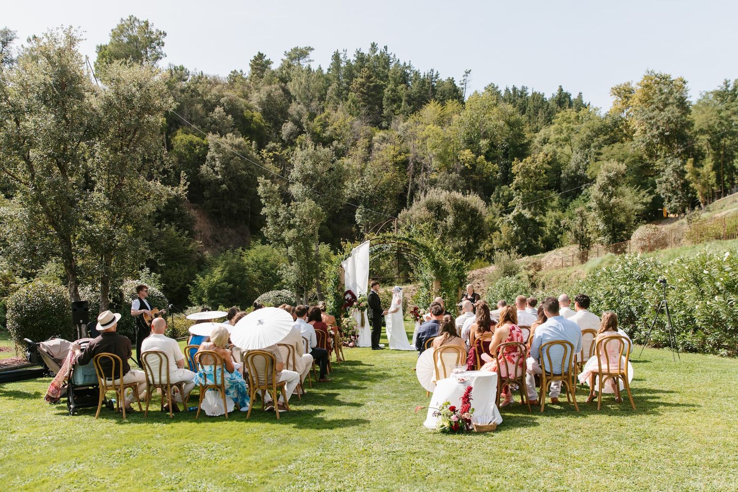 Outdoor wedding ceremony on green lawn with guests seated in wooden chairs facing floral arch, surrounded by lush trees.