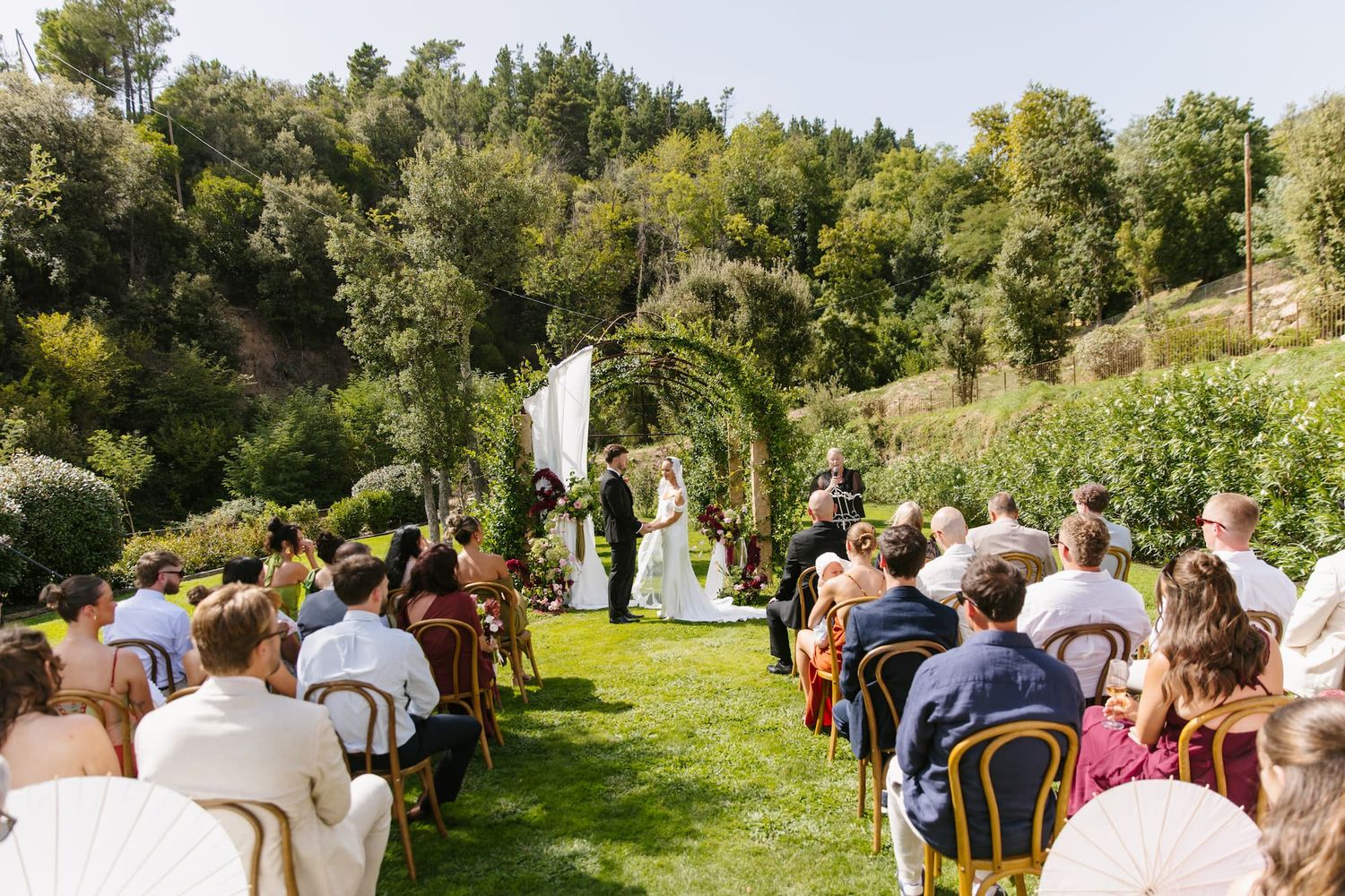 Outdoor wedding ceremony on green lawn with floral arch, seated guests, and lush trees in background.