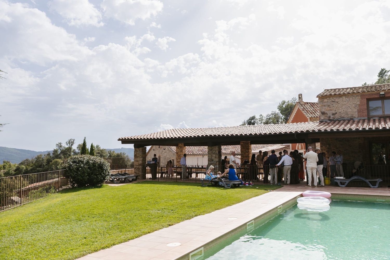 Elegant outdoor villa pool party with guests gathered under a pergola on a sunny day in a Mediterranean setting.