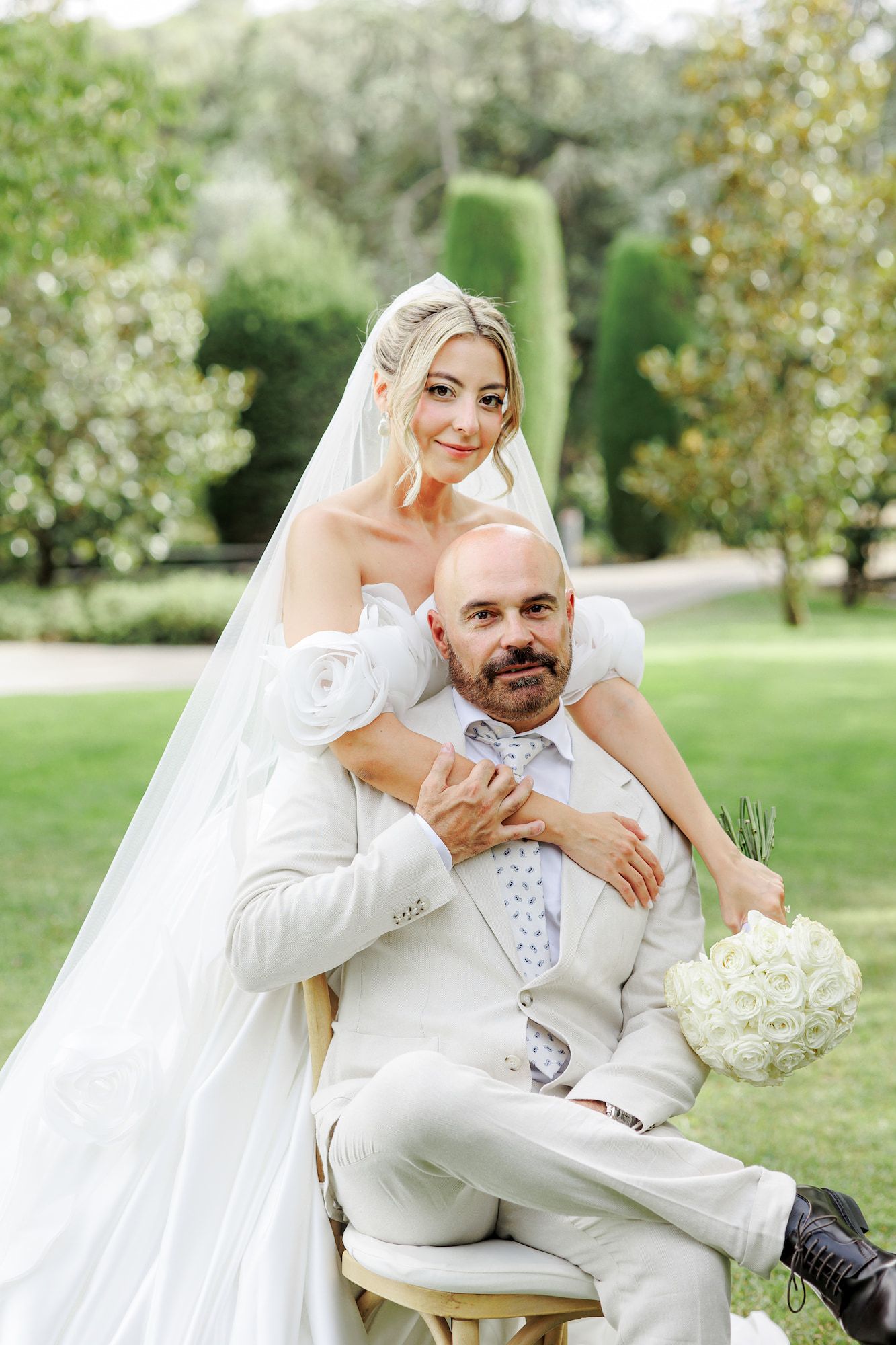 Bride in white gown with veil embraces seated groom in white suit holding bouquet in lush garden setting.