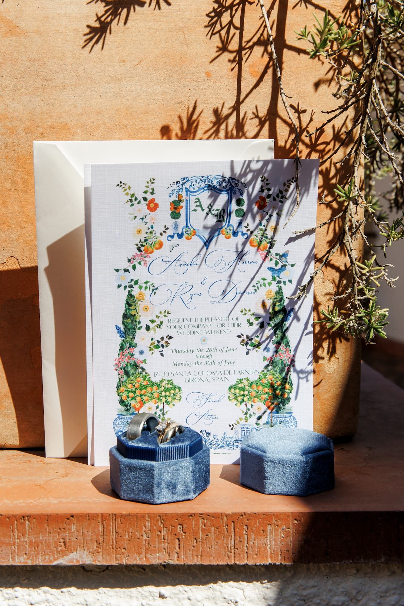 A wedding invitation with a floral design sits in sunlight next to two blue candles on a terracotta surface.