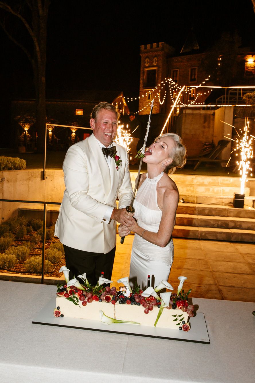 Bride and groom in white laugh while cutting a floral wedding cake with a sparkler at a nighttime outdoor reception with twinkling lights.