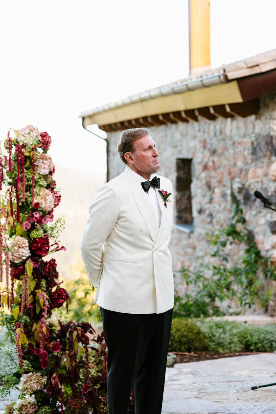 Groom in a white dinner jacket and black bow tie stands near a tall red and pink floral arrangement beside a rustic stone building.