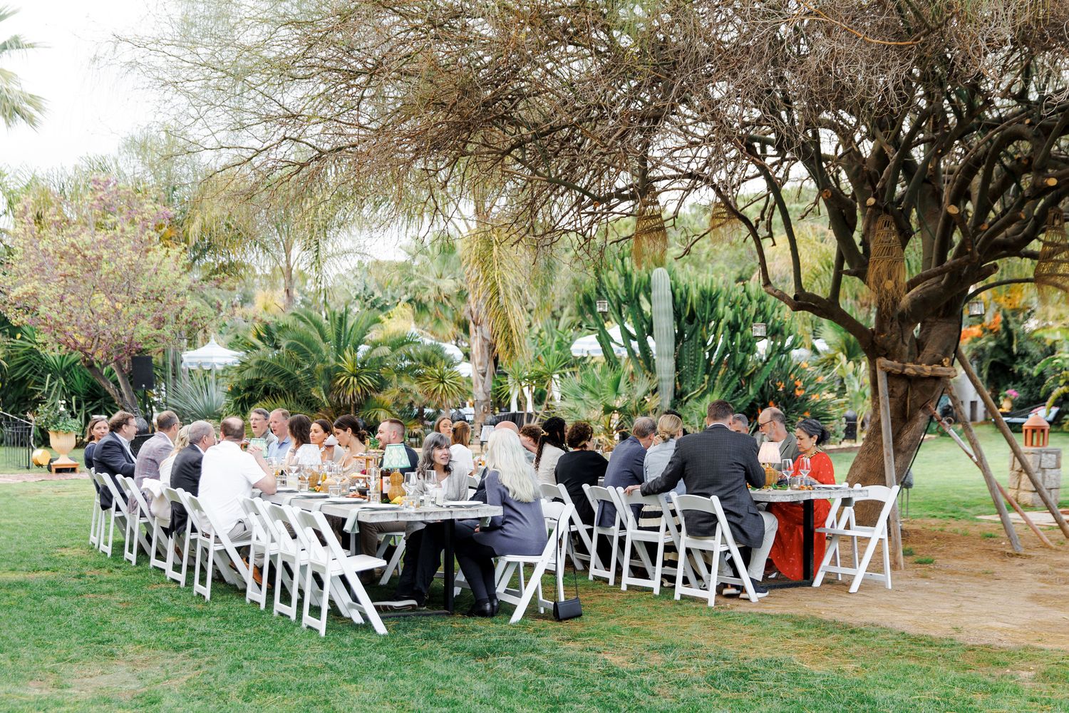 Guests dining outdoors at a long white table under trees in a lush garden setting during a wedding reception.