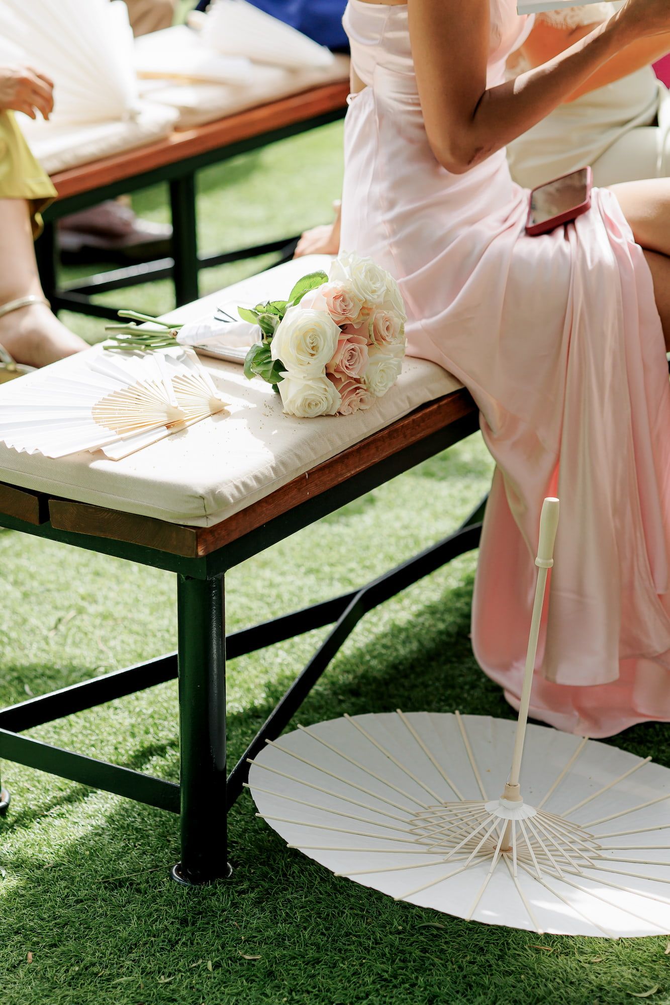 Bridesmaid in pink dress seated on bench with white rose bouquet, hand fans, and parasol at outdoor wedding.