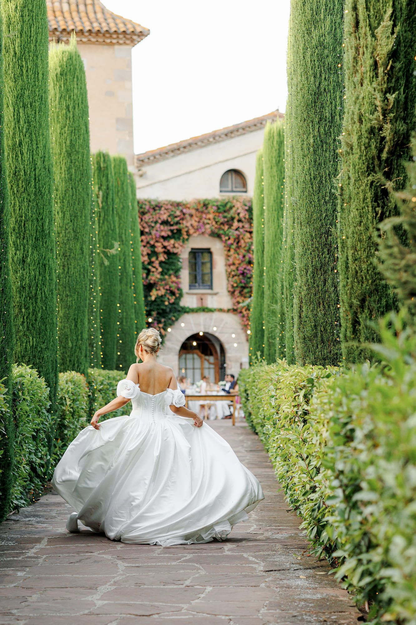 Bride in a white ball gown walks through a lush garden pathway lined with tall cypress trees toward a vine-covered villa.