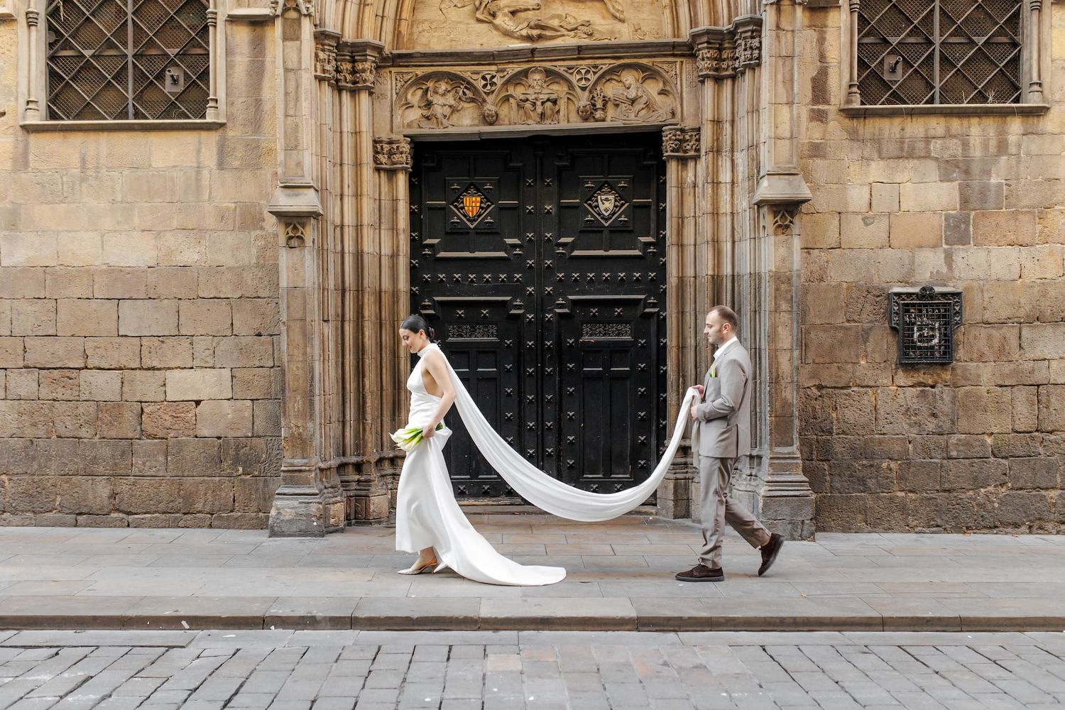 A bride in a flowing white dress walks along a historic stone building facade with ornate black doors and windows.
