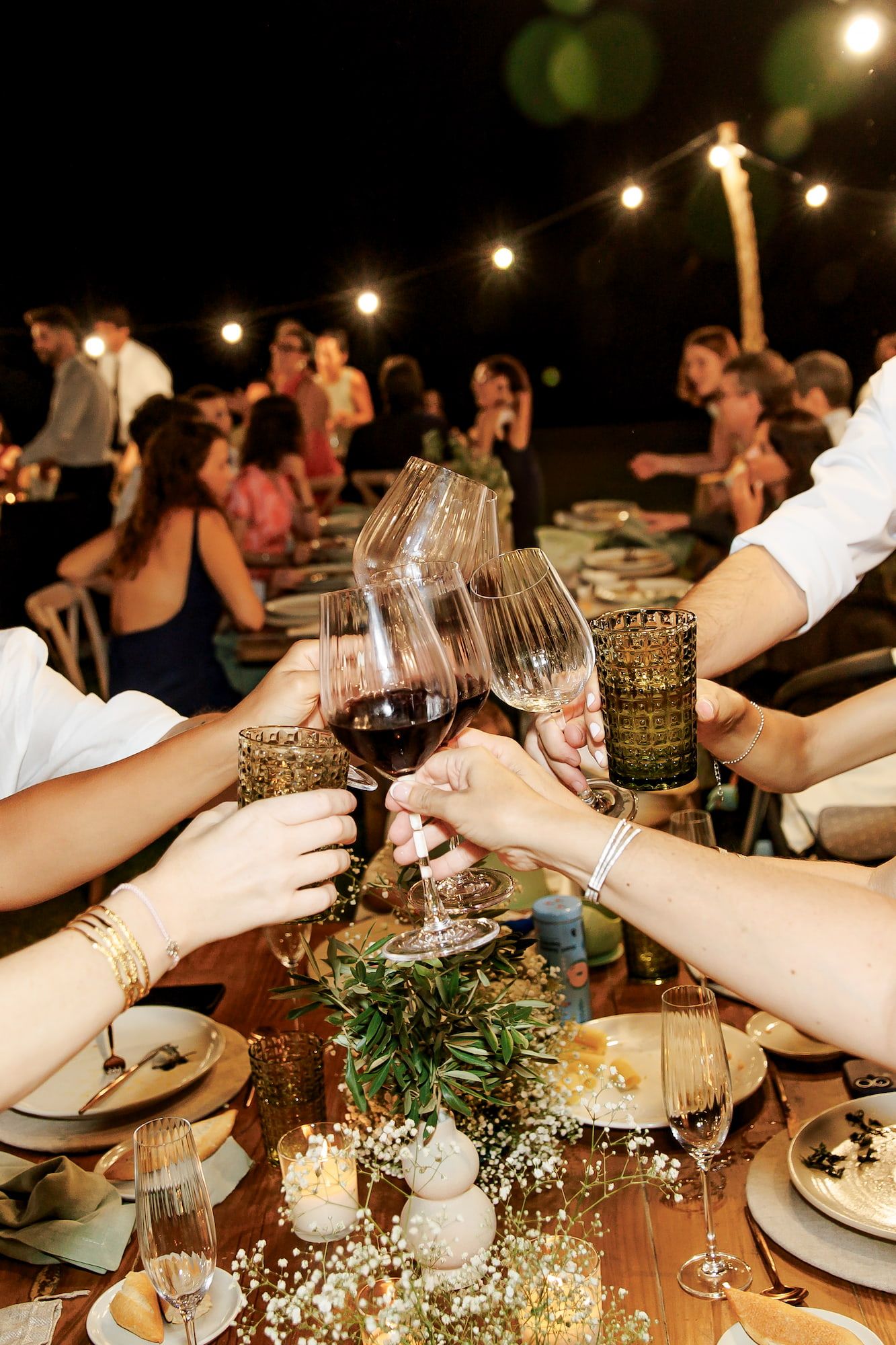 Guests clinking wine glasses in a toast at an elegant outdoor dinner party with string lights at night.