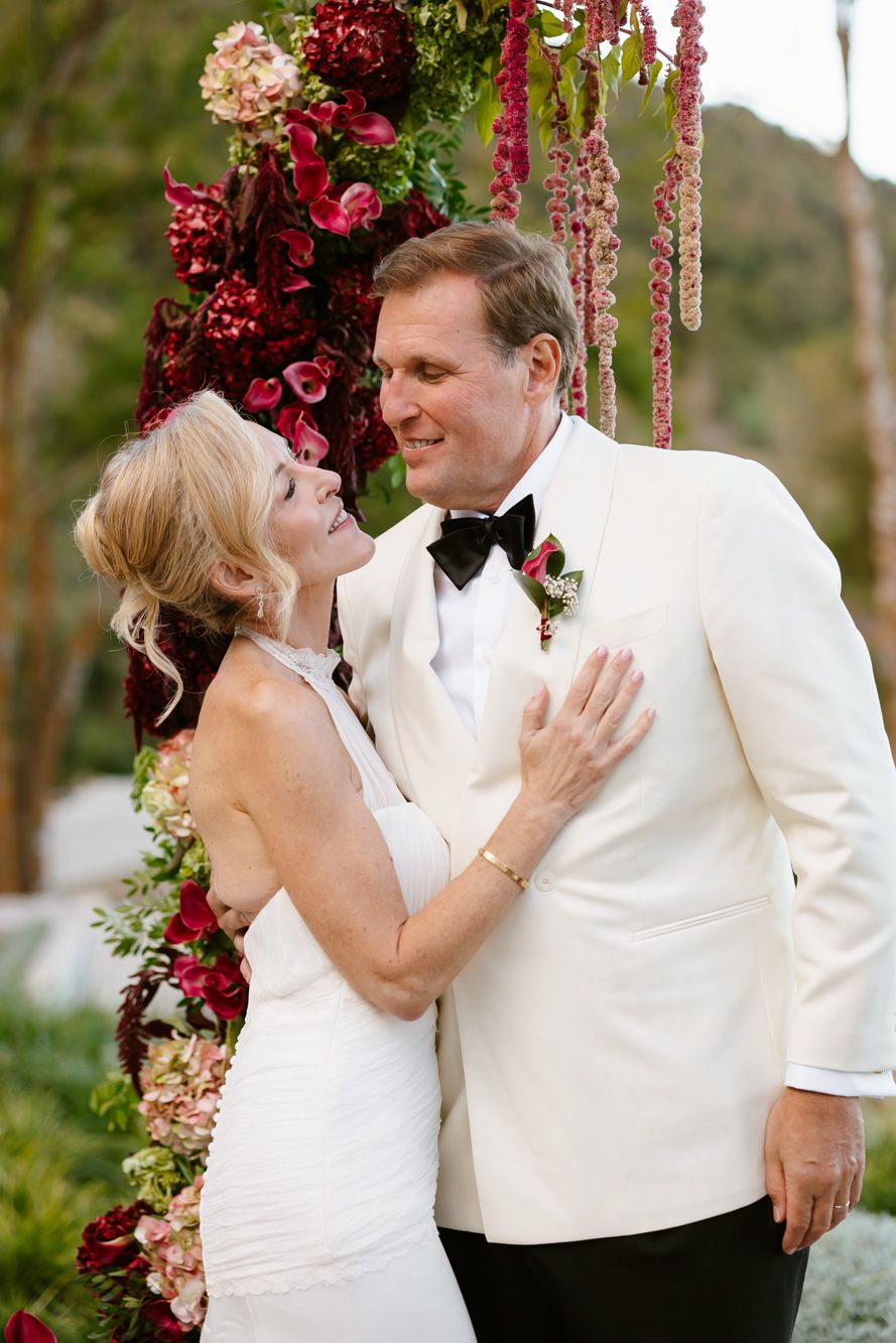 Bride in a white halter gown and groom in a white dinner jacket and black bow tie embrace beneath a lush red and pink floral wedding arch.