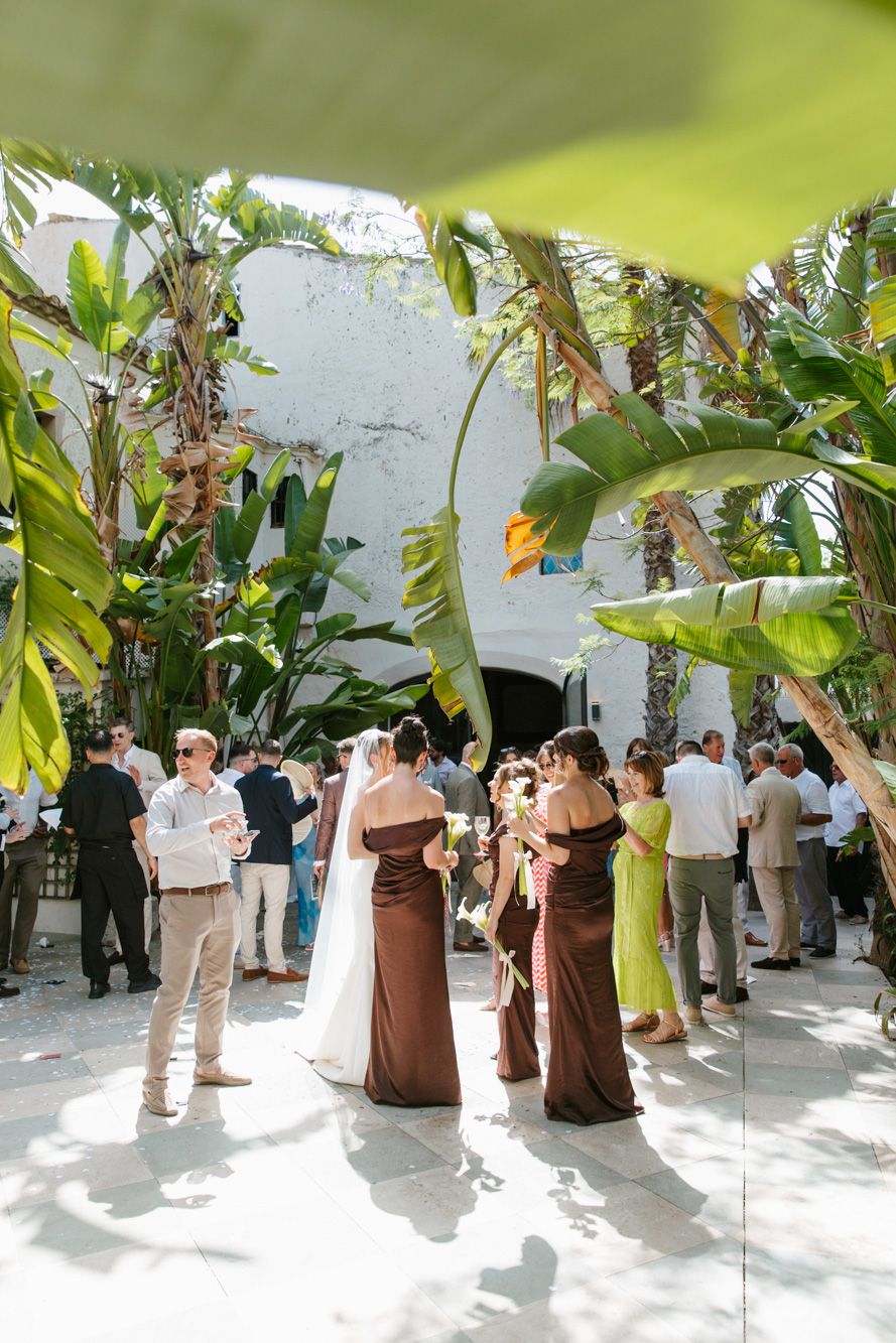 Bride and bridesmaids in brown and green dresses gather in a tropical courtyard during an outdoor wedding ceremony.