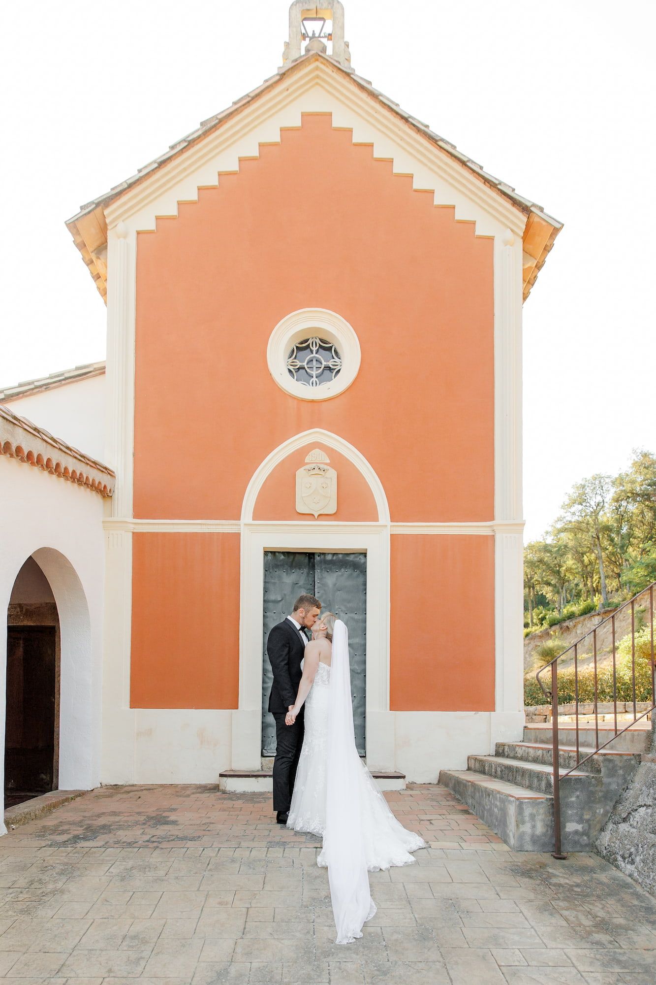 A couple shares romantic moments outside of a peach-colored Spanish church with white trim.