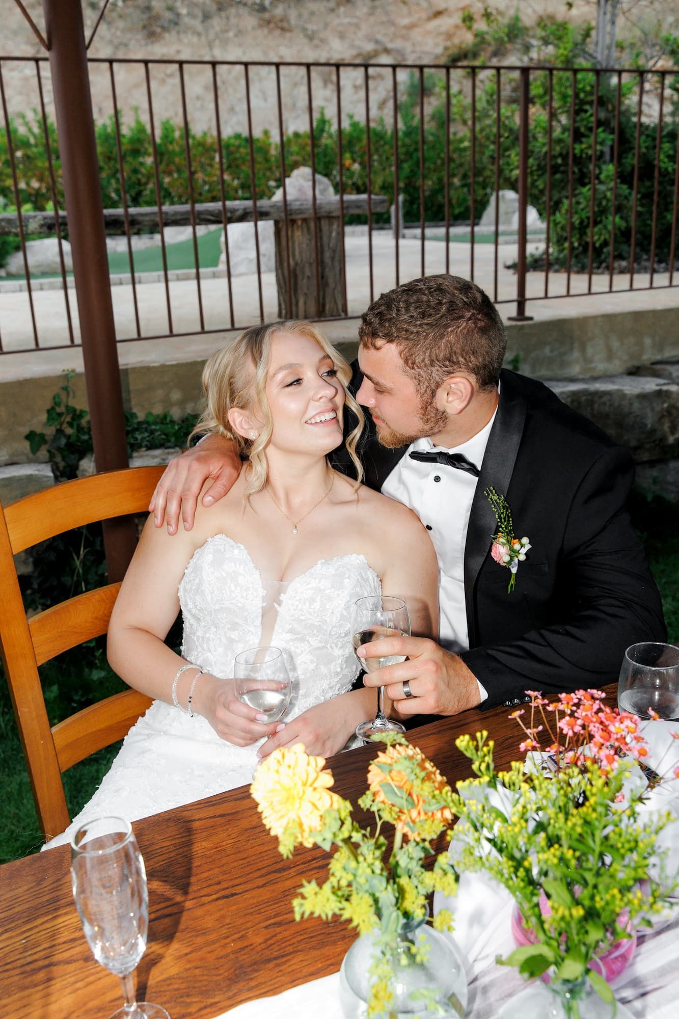 A couple in wedding attire shares an intimate moment at a table decorated with floral centerpieces.