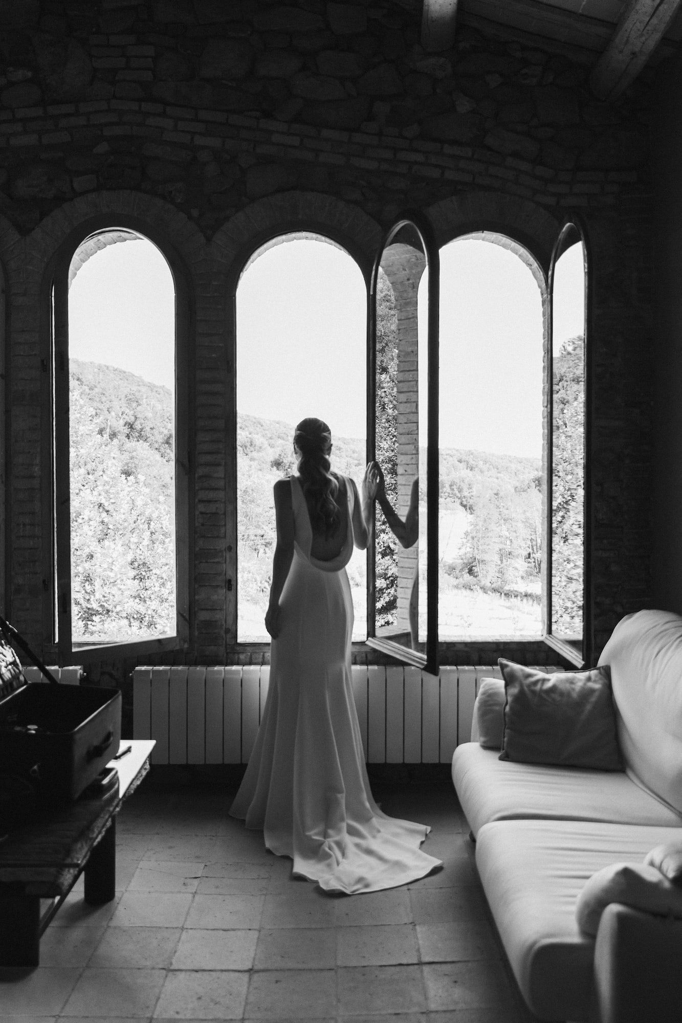 Bride in elegant backless wedding dress stands before arched windows overlooking lush hills in black and white photo.