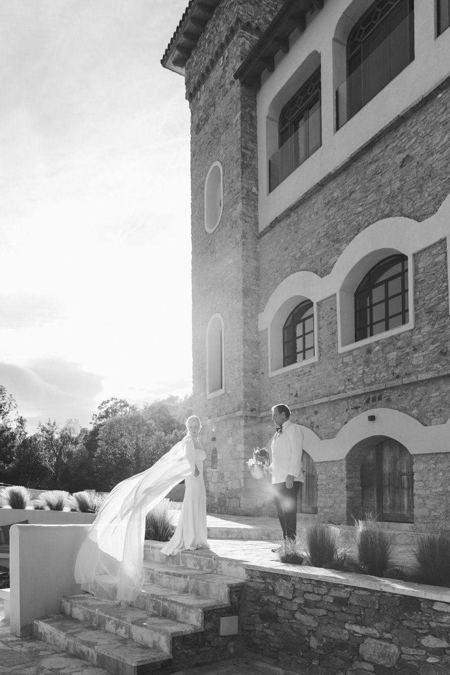 Black and white photo of a bride in a flowing veil and a groom in shirtsleeves on stone steps beside a historic arched stone building.