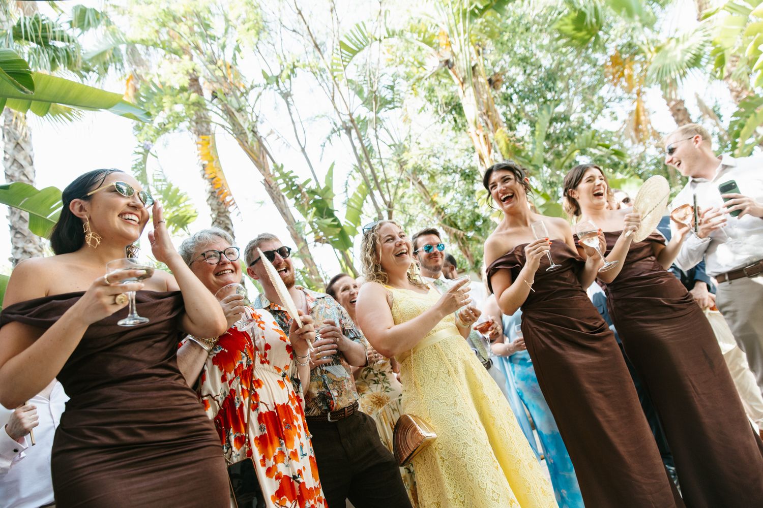 Bridesmaids in brown strapless gowns laugh and celebrate outdoors at a tropical wedding reception.