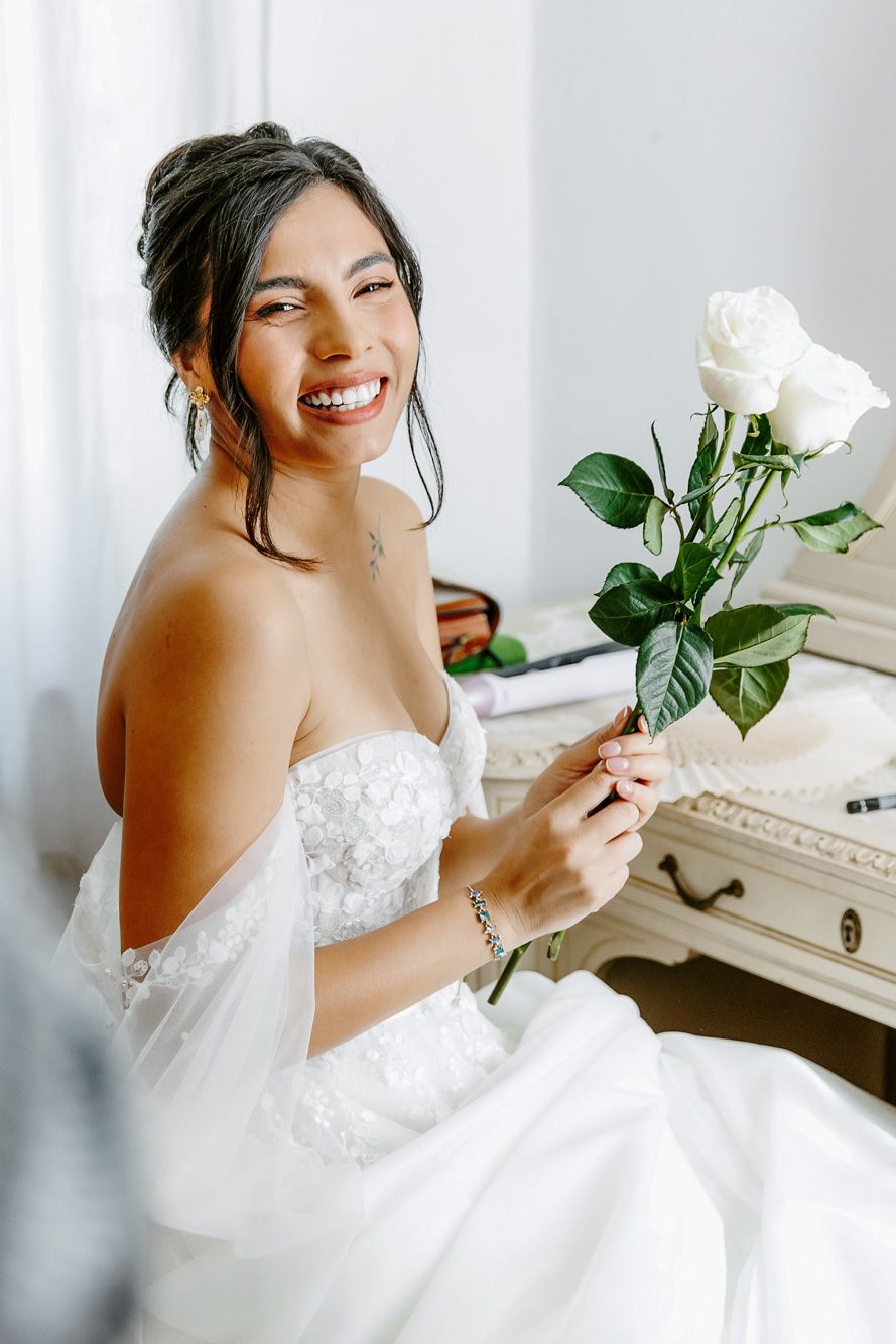 Smiling bride in strapless white lace gown holding white roses, wearing diamond jewelry, seated by vintage dresser.