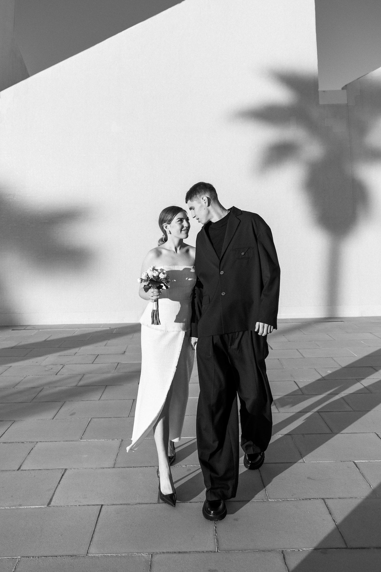 Black and white photo of a bride and groom posing against a white wall with a palm tree shadow.