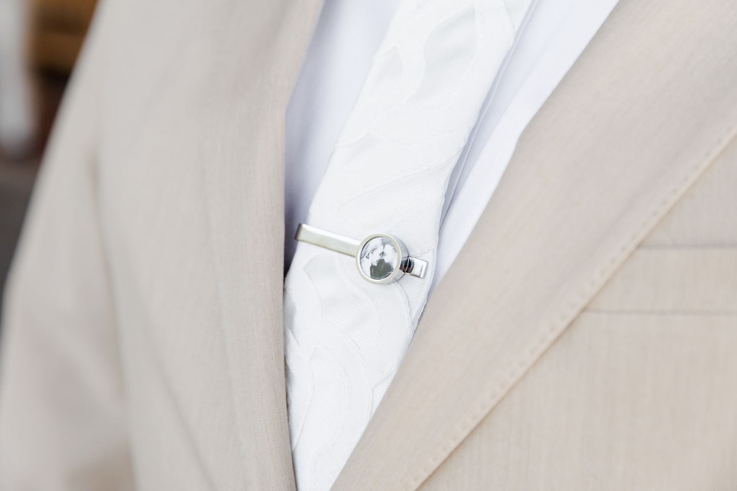 Close-up of silver tie pin on white dress shirt under beige wedding suit jacket.