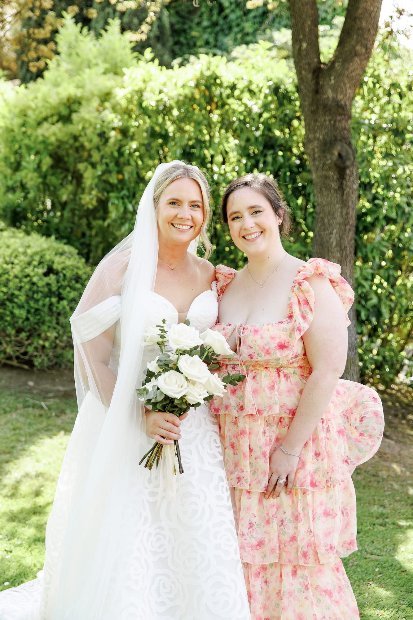 Bride in white gown holding bouquet poses with bridesmaid in pink floral dress outdoors.