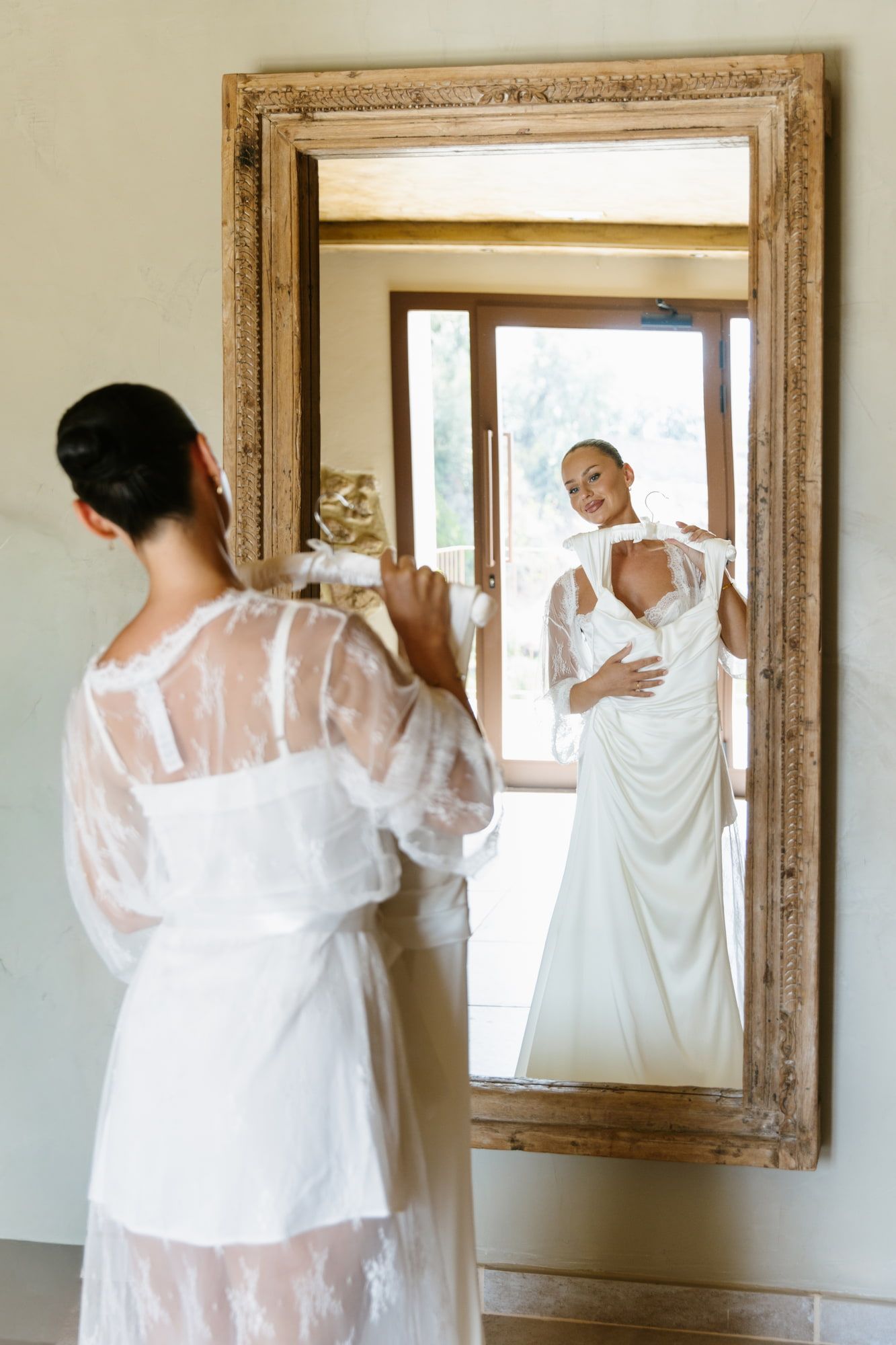 Bride in sheer white robe holds wedding dress while smiling at her reflection in an ornate wooden mirror.