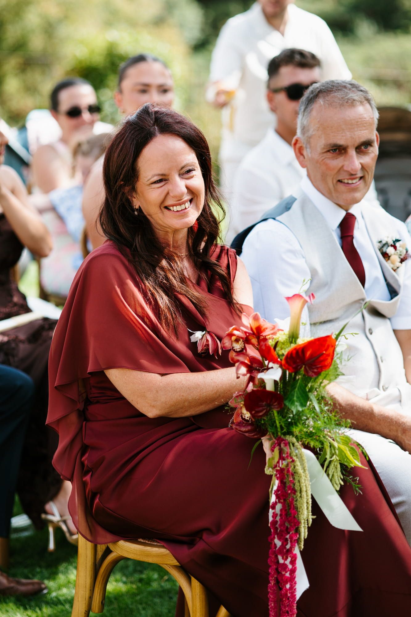 Smiling woman in burgundy dress holding red floral bouquet seated at outdoor wedding ceremony.