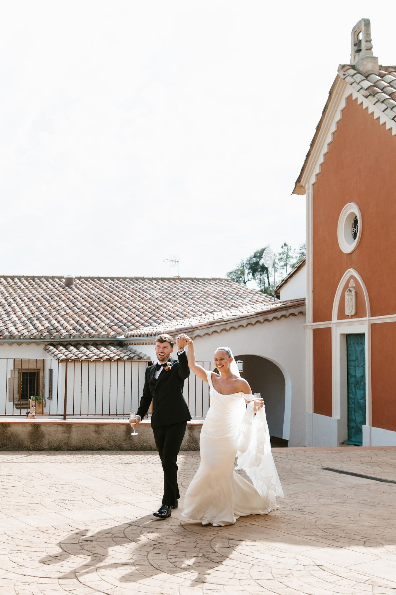 Bride and groom dancing joyfully in a sunlit courtyard beside a charming red Mediterranean chapel on their wedding day.