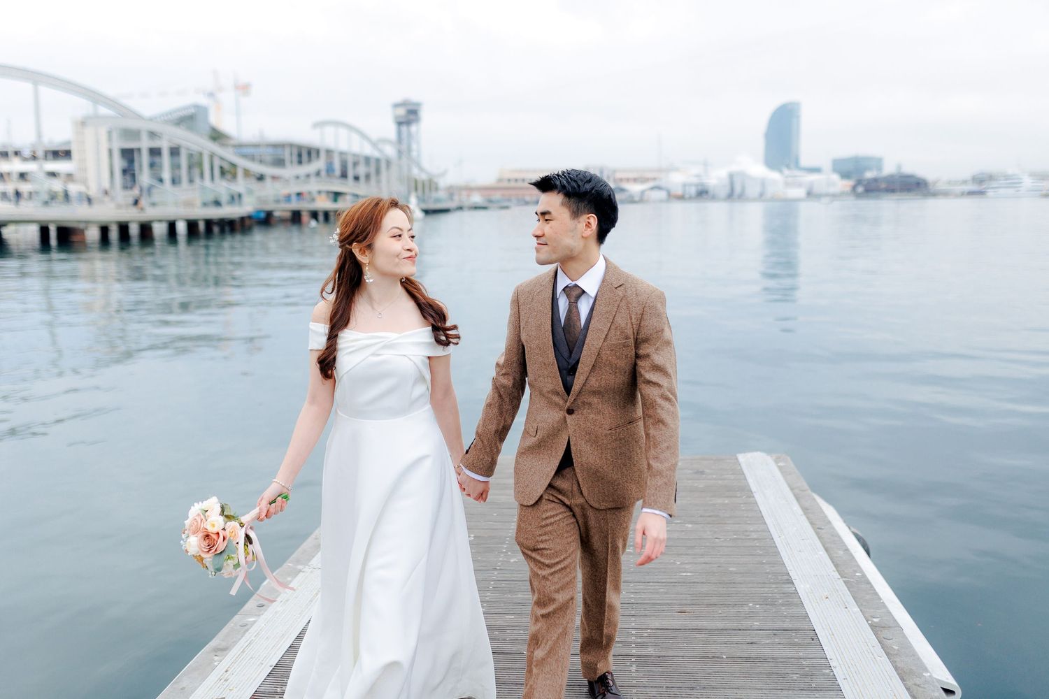 A newly married couple walks down  the port Vell in Barcelona and look over the deep blue water.