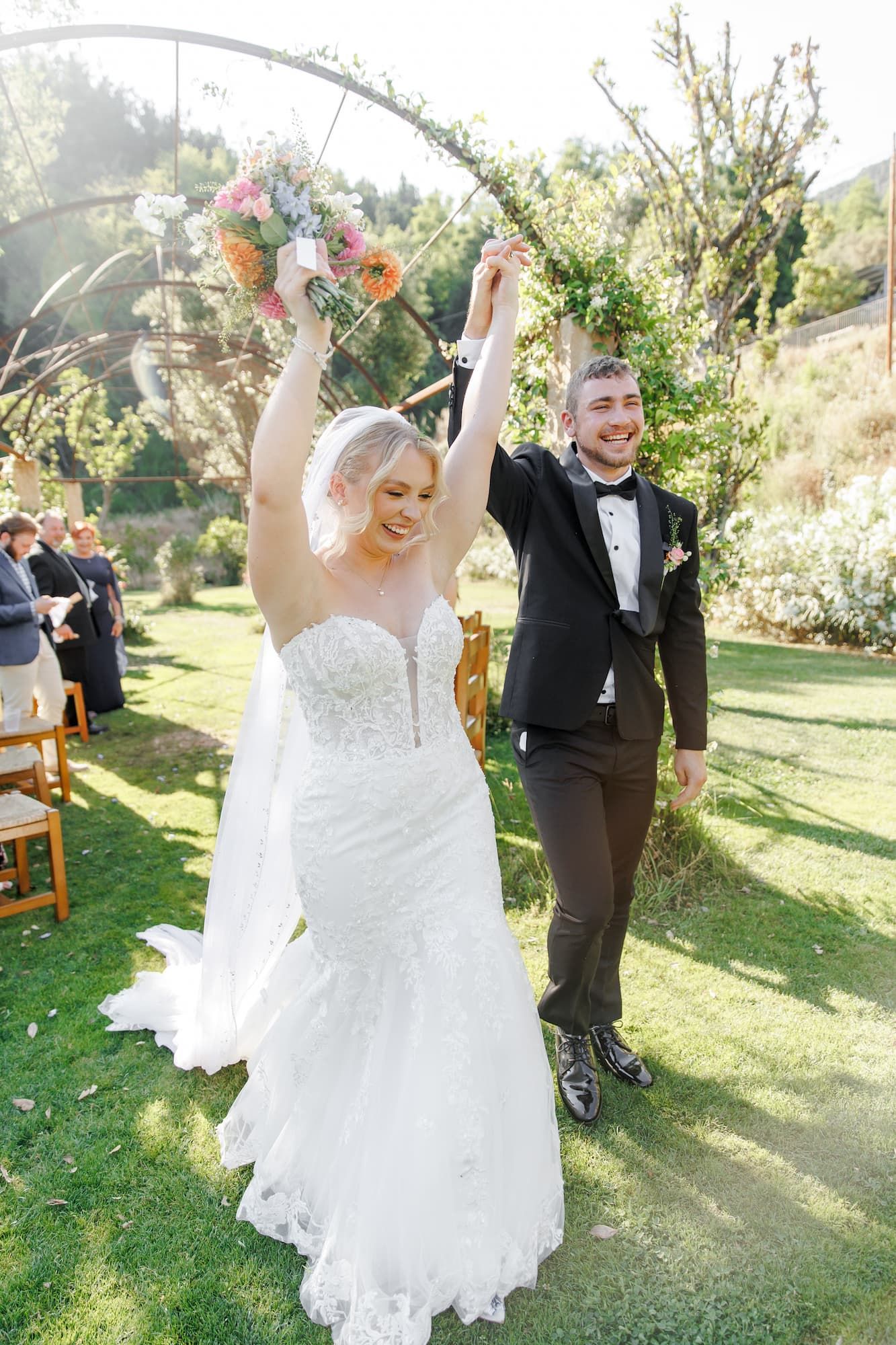 A joyful couple celebrates during an outdoor garden wedding ceremony, with guests watching in natural sunlight.