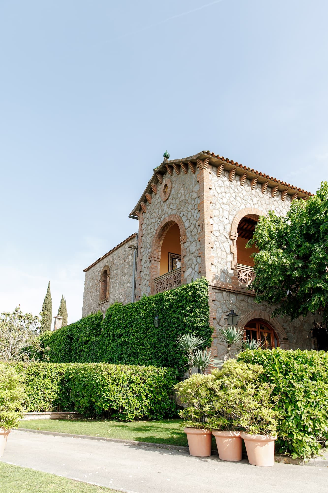 A stunning Mas de Sant Lleí villa with arched windows, lush green hedges, and terracotta pots under a clear blue sky.