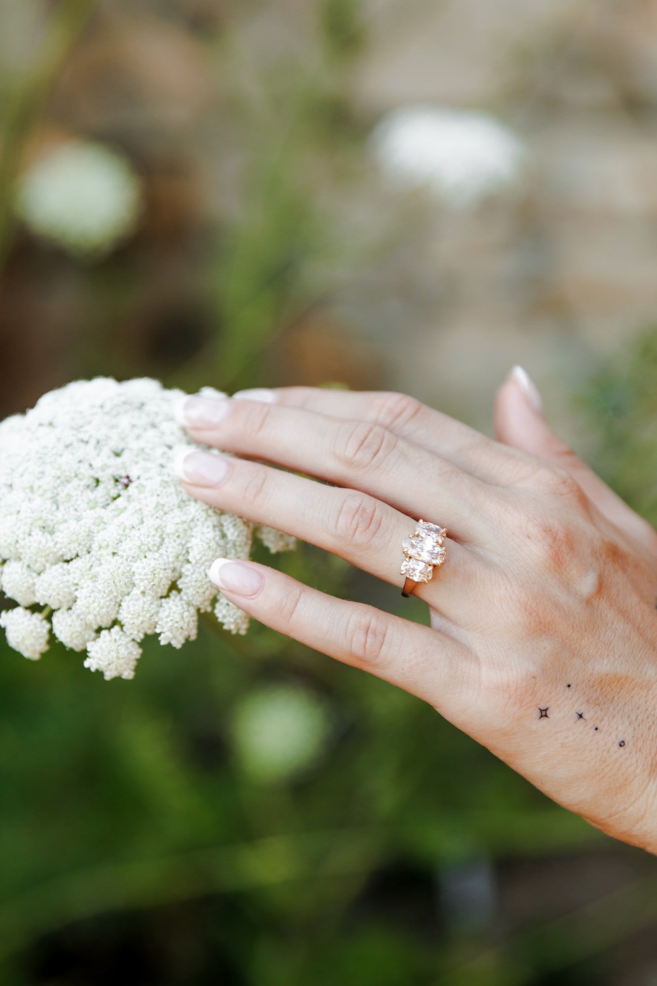 Close-up of a hand wearing a rose gold diamond engagement ring, gently touching white Queen Anne's Lace flowers.