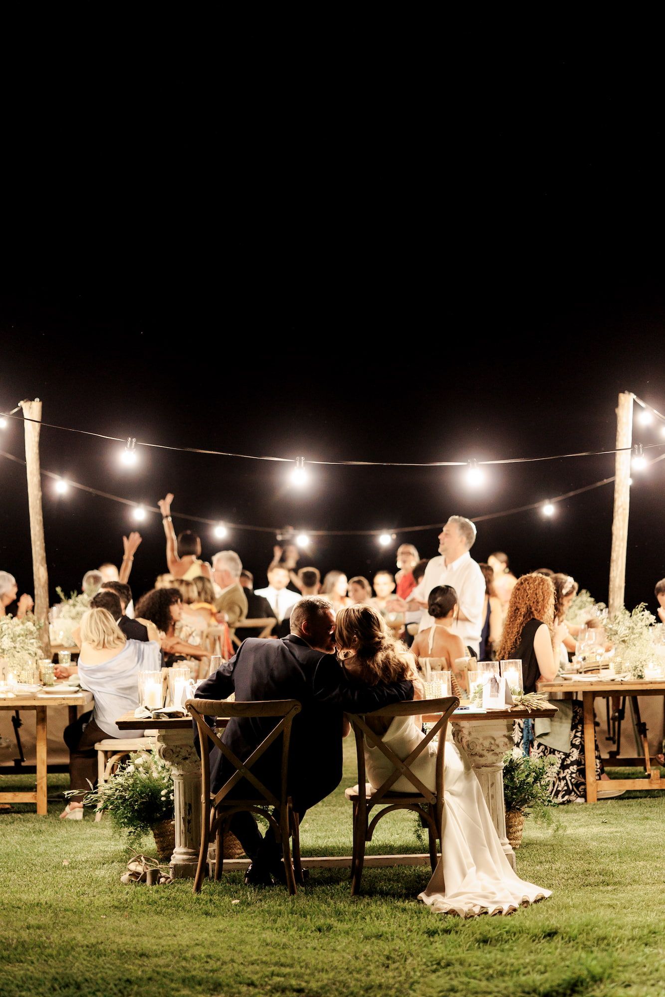 Romantic outdoor wedding reception at night with string lights, guests gathered around rustic wooden tables on a green lawn.