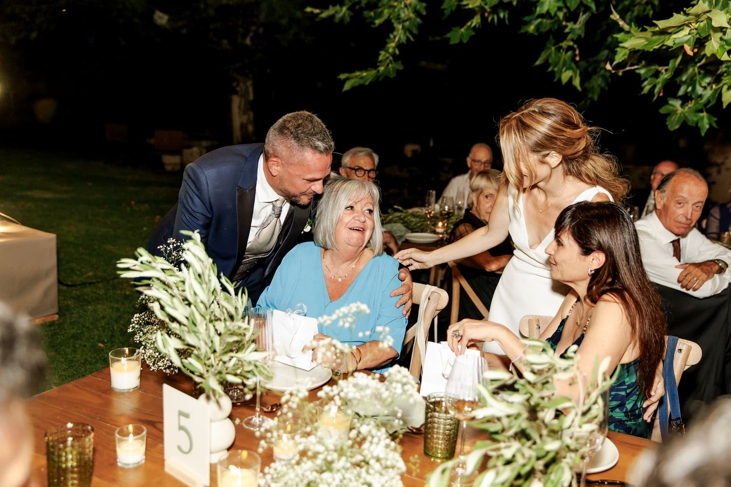 Bride and groom greeting elderly guest at outdoor wedding reception table decorated with greenery and candles.