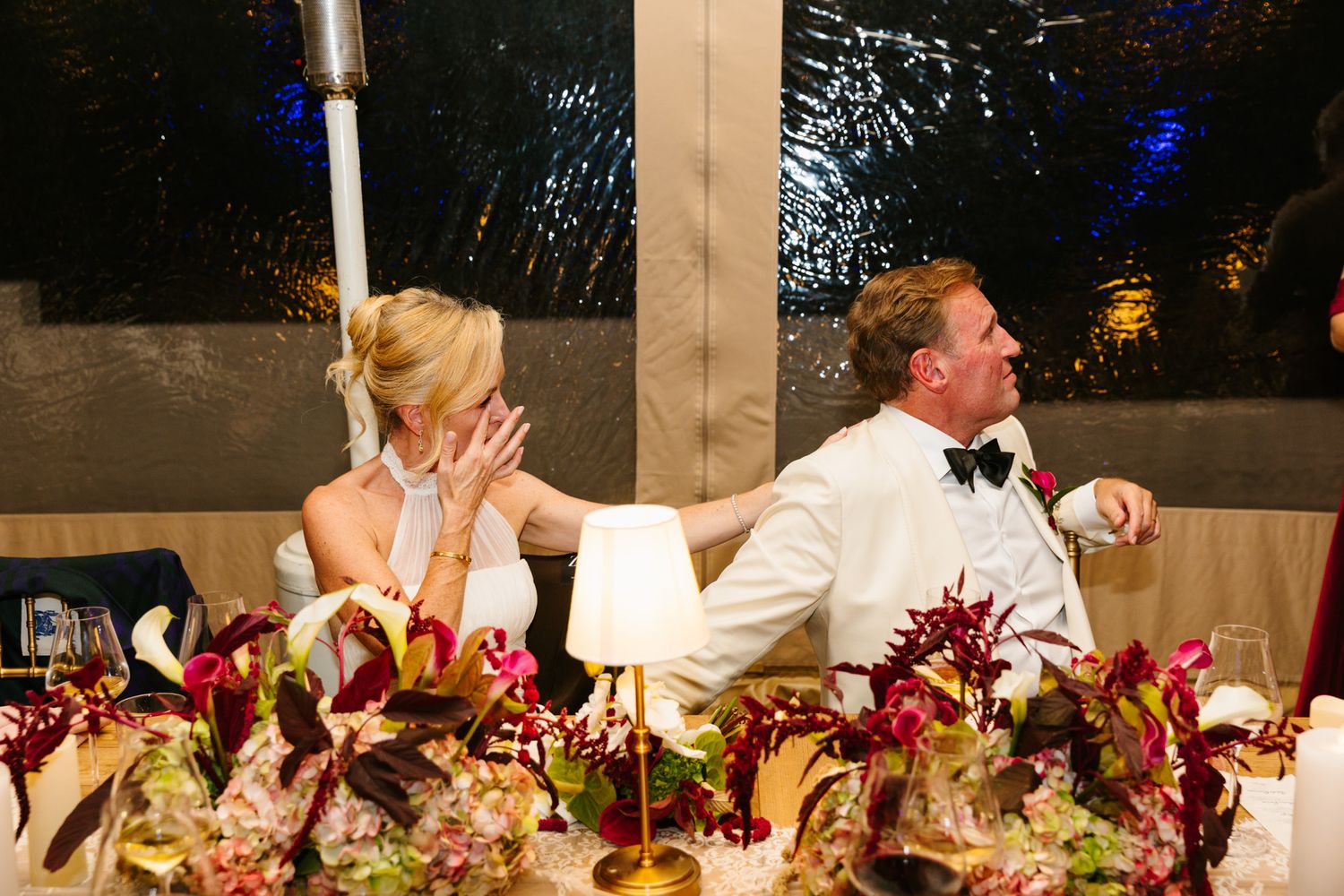 Bride in a white dress wipes her eyes beside a groom in a white jacket and bow tie at a reception table with colorful floral centerpieces.