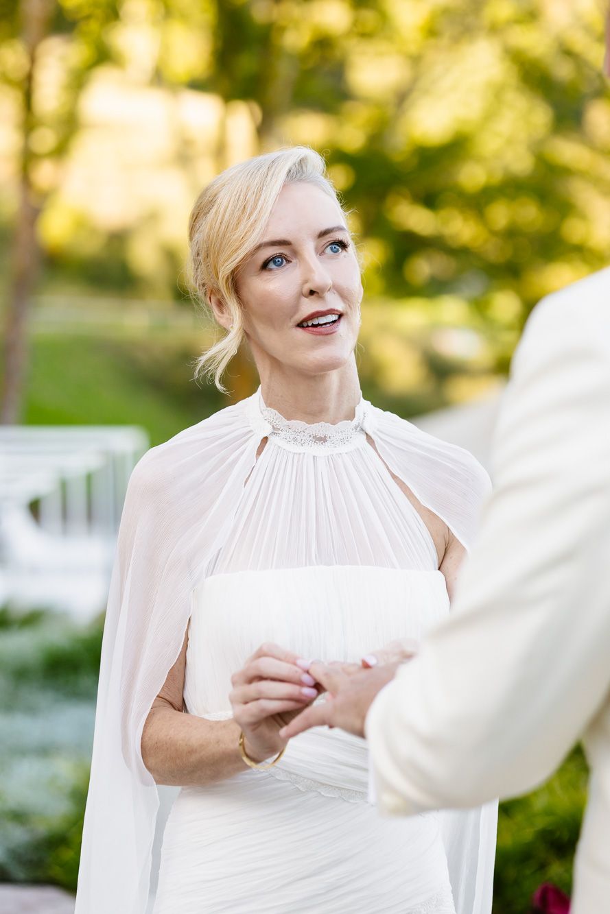 Bride in a white gown with sheer cape holds hands with her groom during an outdoor wedding ceremony surrounded by greenery.