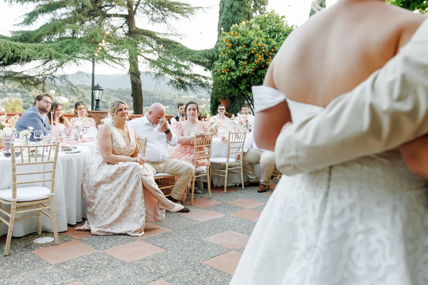 Elegant outdoor wedding reception with guests seated at gold chiavari chairs, bride in off-shoulder gown in foreground.
