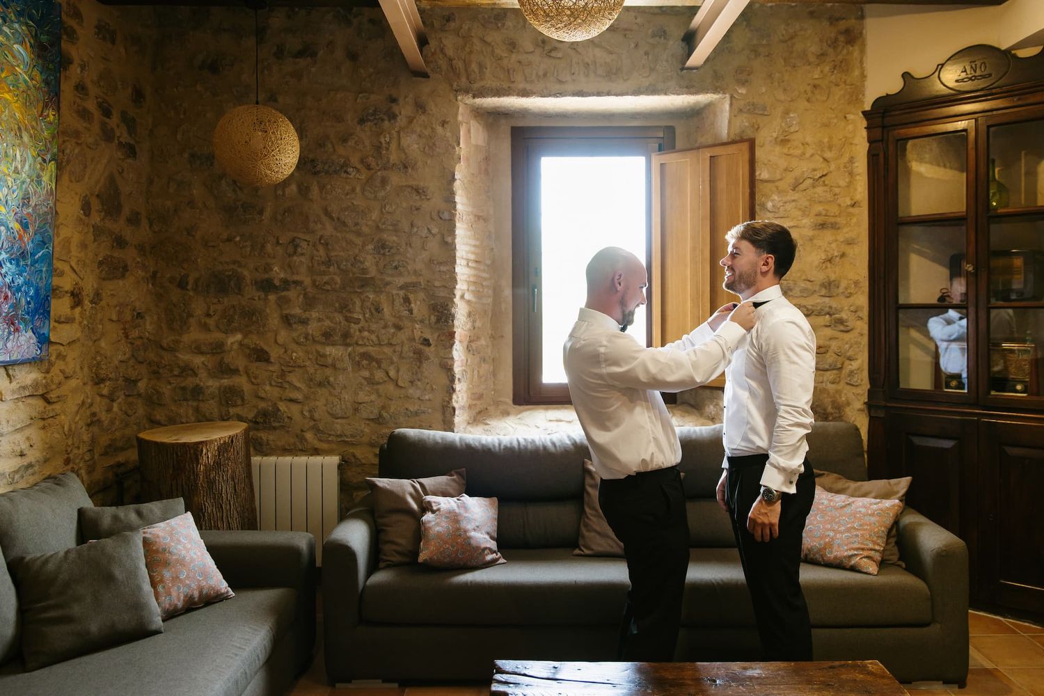 A man helps a groom adjust his tie in a rustic stone-walled room before a wedding ceremony.