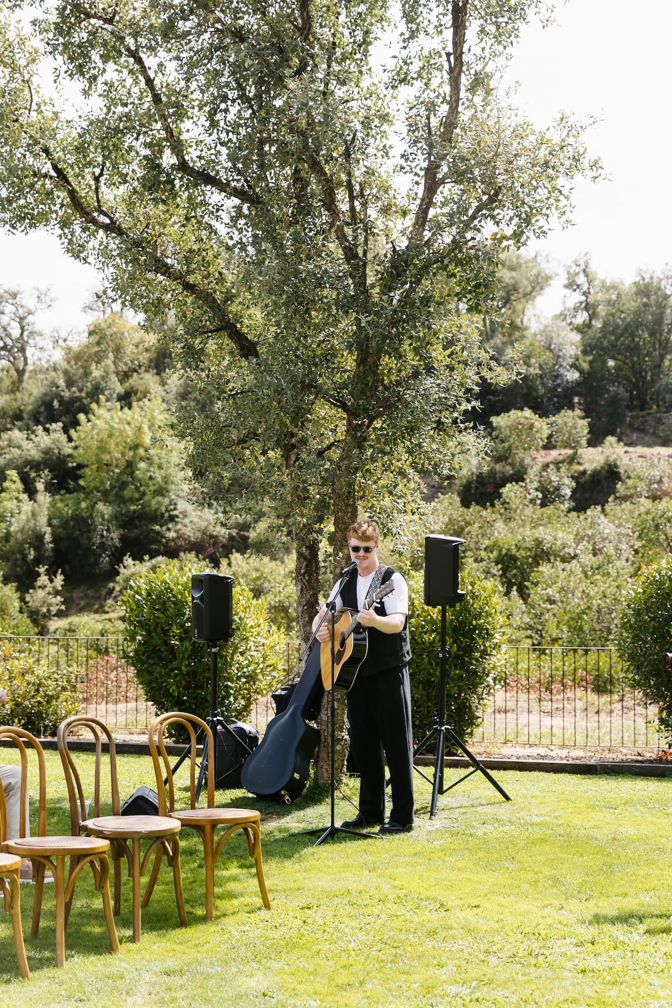 Musician playing acoustic guitar outdoors at a garden wedding ceremony with speakers and wooden chairs on green lawn.