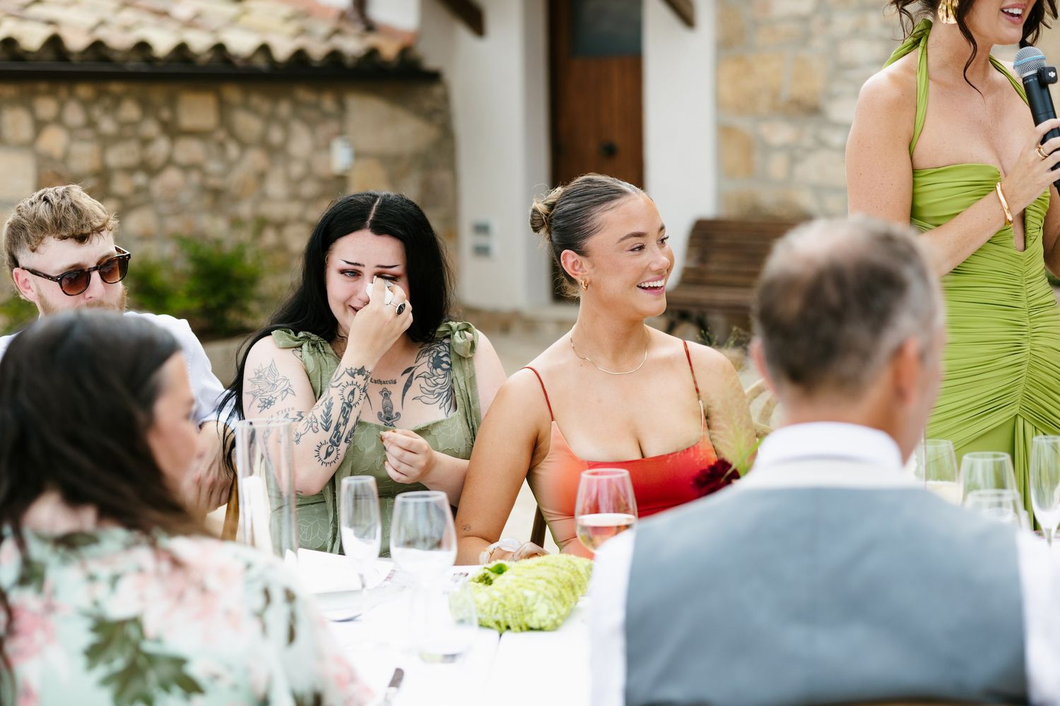 Guests laughing and crying happy tears at an outdoor wedding reception dinner table in a rustic venue.