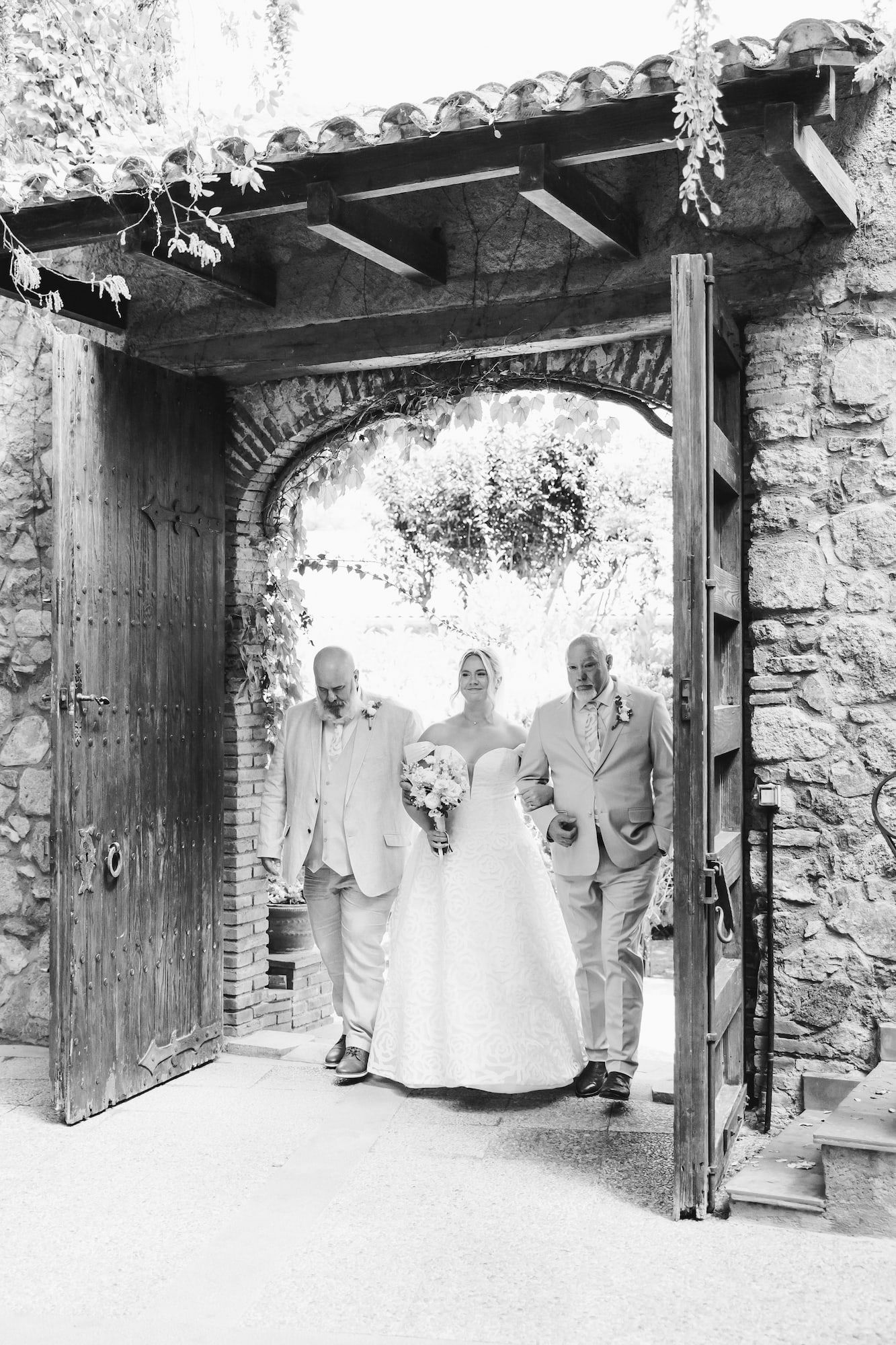 Bride in white gown walks through stone arch with two escorts at elegant outdoor wedding venue.