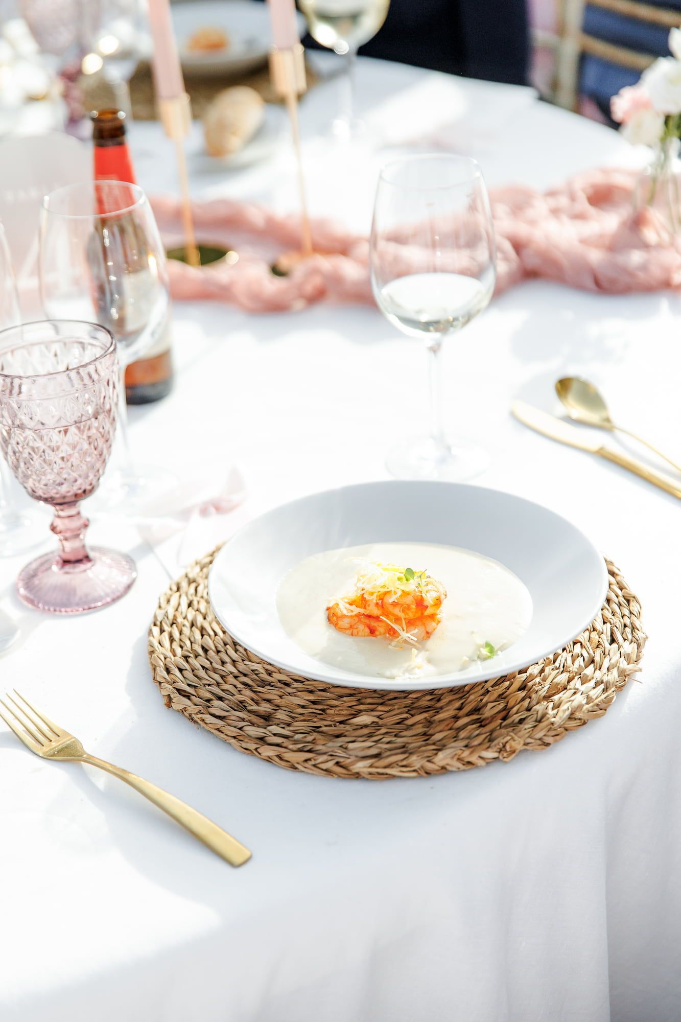 Elegant wedding table setting with cream soup, gold cutlery, pink glassware, and woven placemat on white linen.