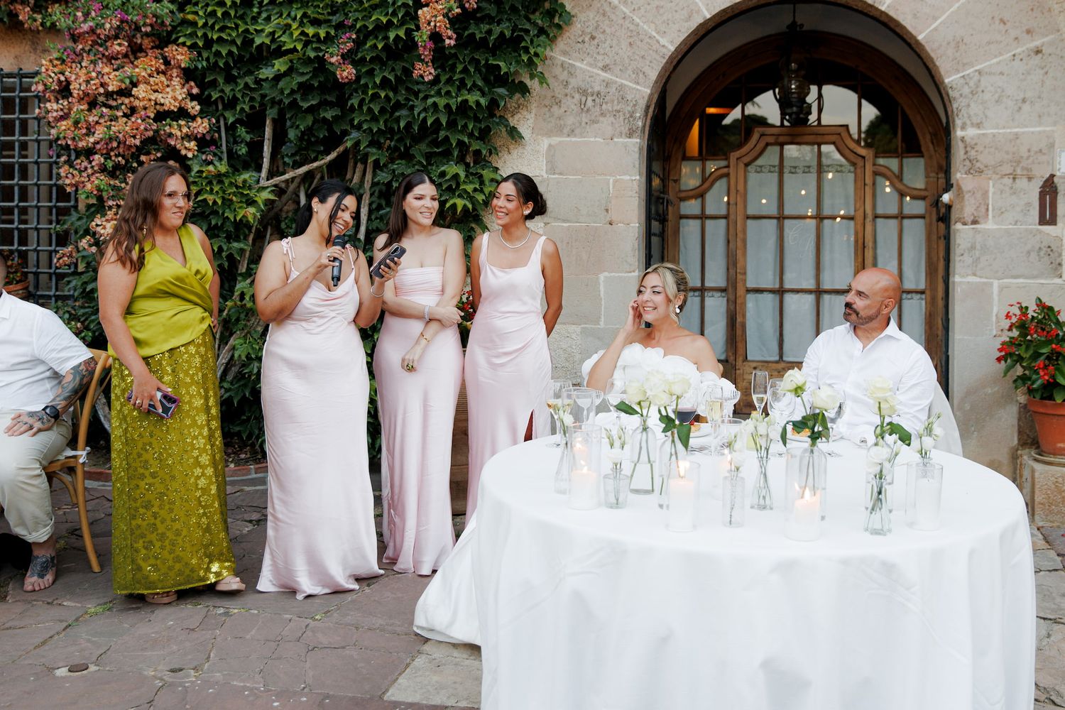 Bridesmaids in pink dresses stand smiling at a wedding reception while bride and groom sit at white floral table.