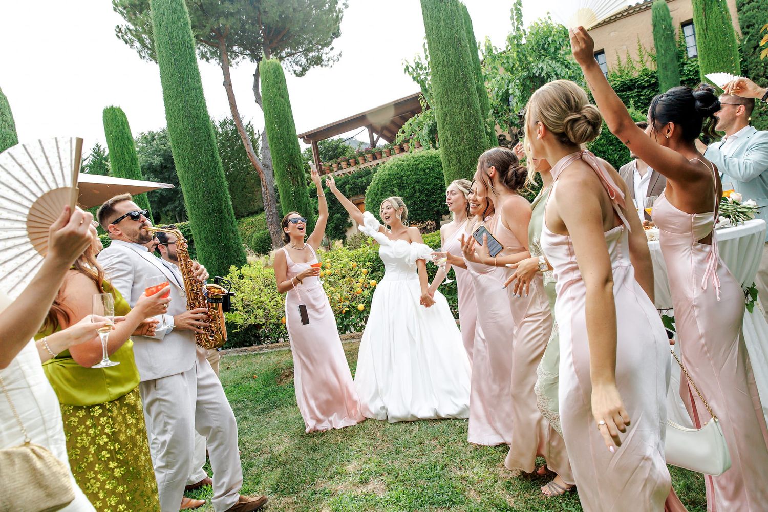 Bride in white gown celebrates with bridesmaids in pink dresses and guests holding fans at outdoor wedding.