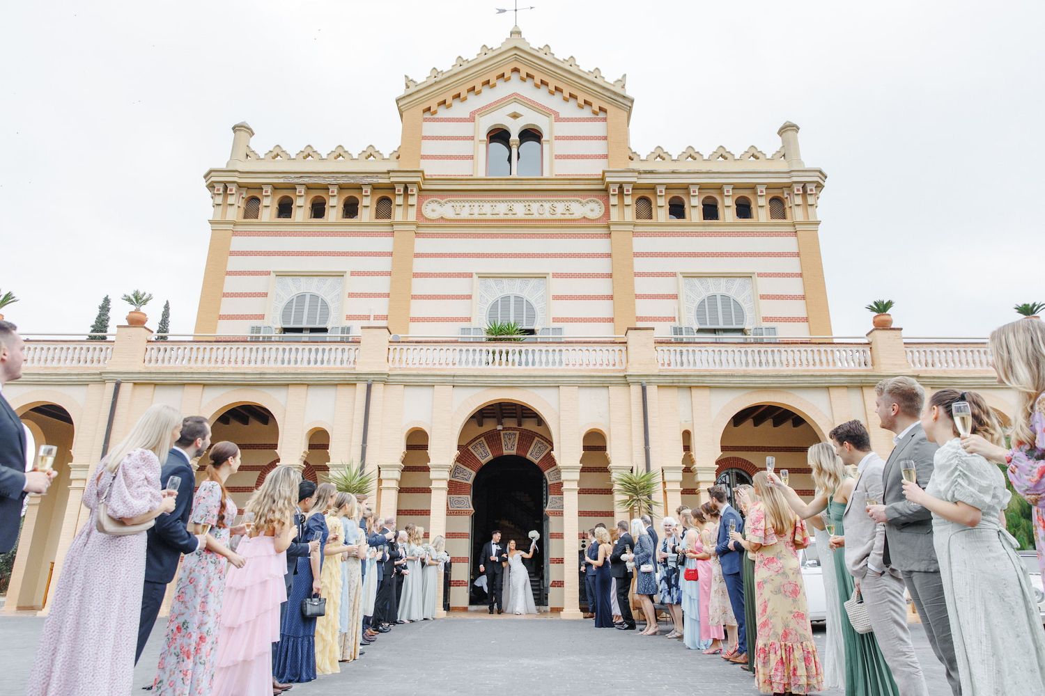 Elegant wedding celebration outside a grand beige Mediterranean-style villa with ornate architectural details.