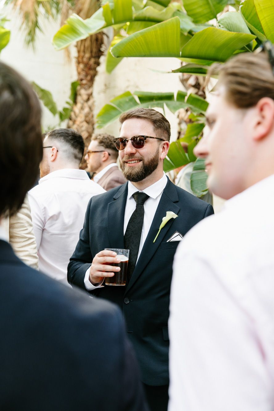 Smiling bearded groom in dark suit and sunglasses holding a beer at an outdoor wedding reception.