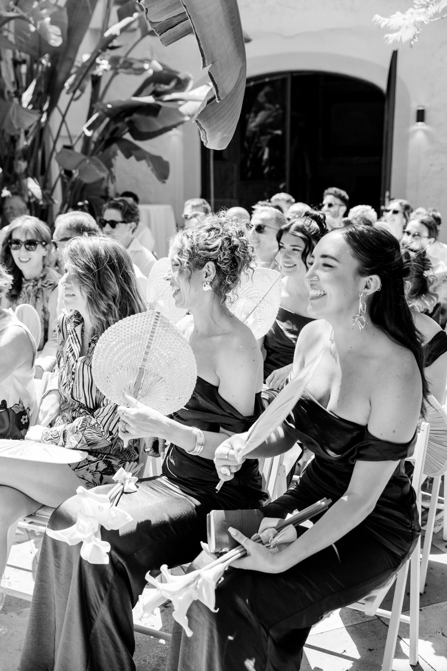 Guests in black dresses fan themselves at an outdoor wedding ceremony, smiling in black and white photo.
