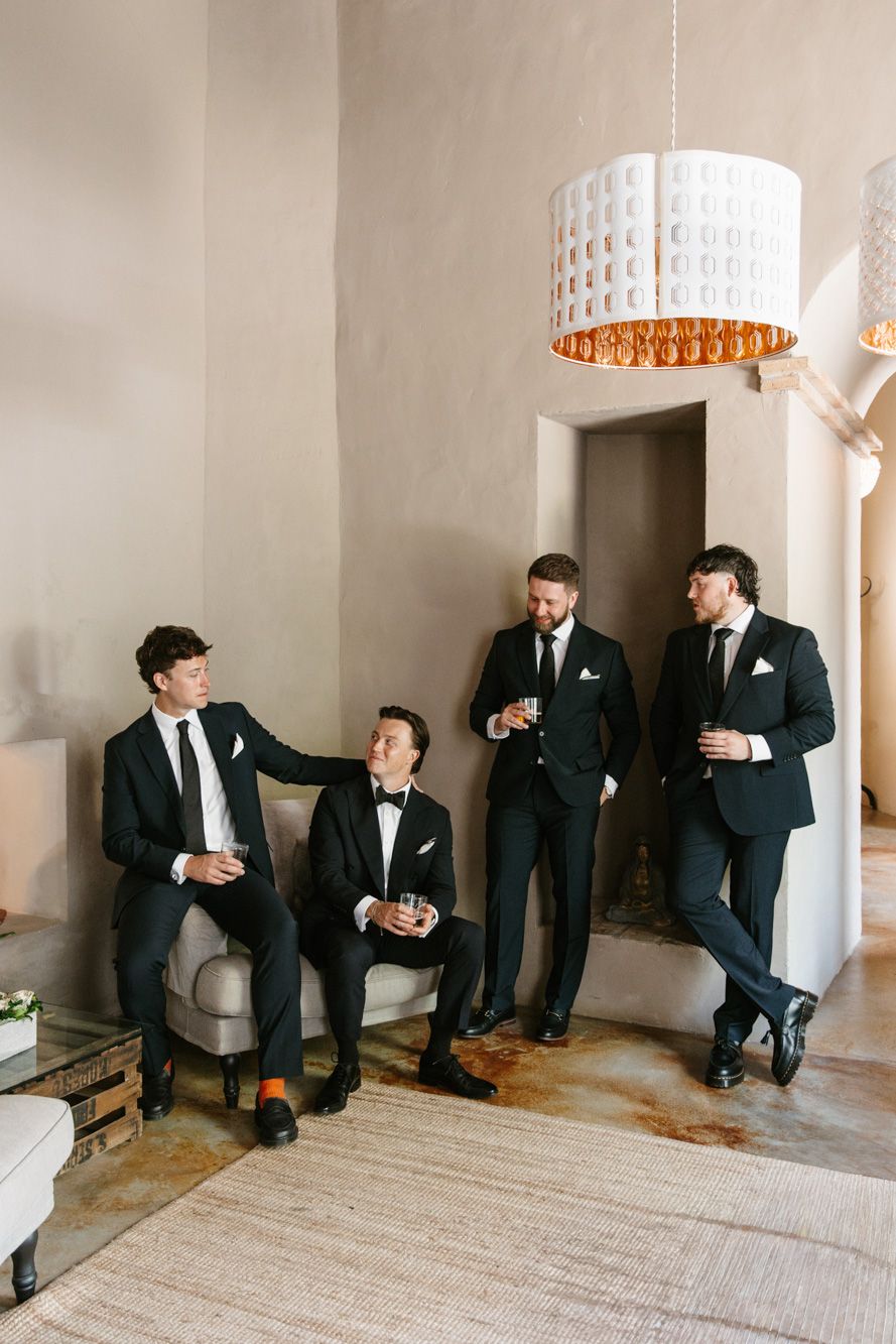 Four groomsmen in black suits socializing with drinks in an elegant neutral-toned room with a decorative chandelier.