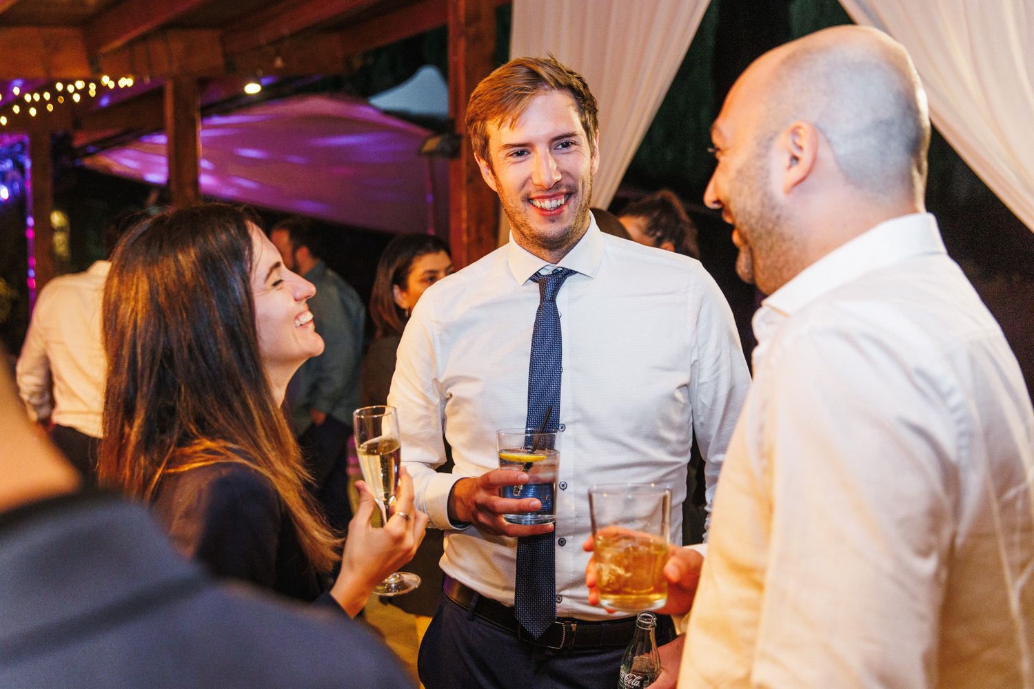 Three guests socializing at a wedding reception, holding drinks and smiling in elegant evening attire.