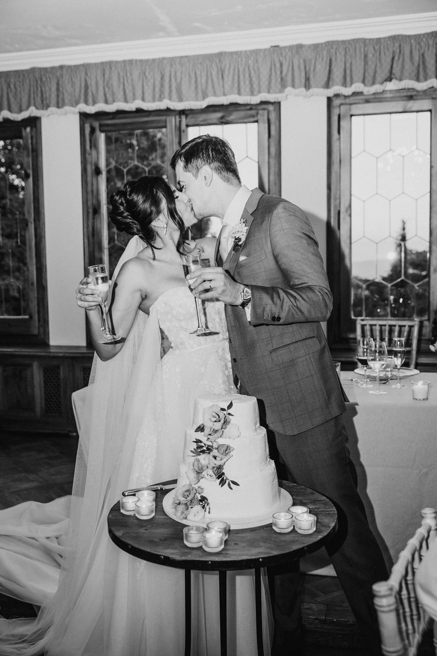 Black and white photo of bride and groom kissing while holding champagne glasses beside a tiered wedding cake at La Baronia
